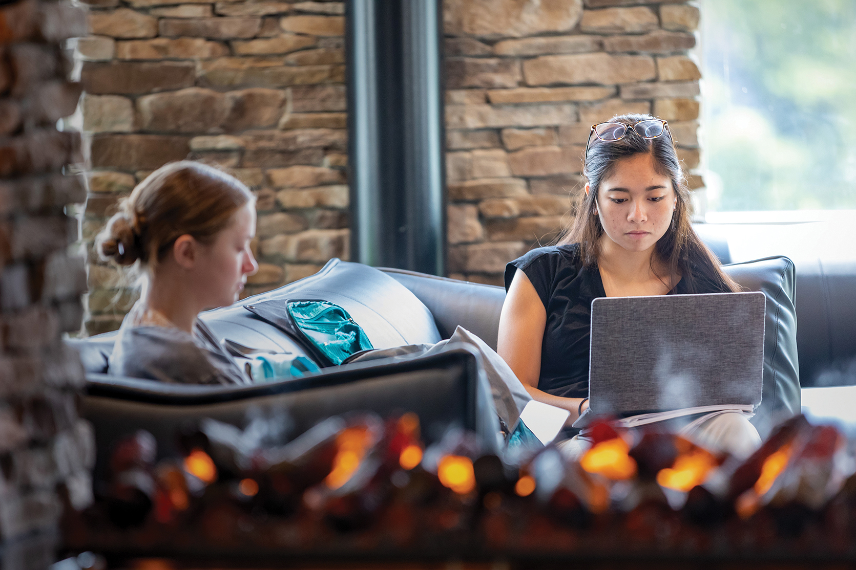 Two women are seated on a dark brown leather couch one is looking at her computer, the other is looking down reading. 